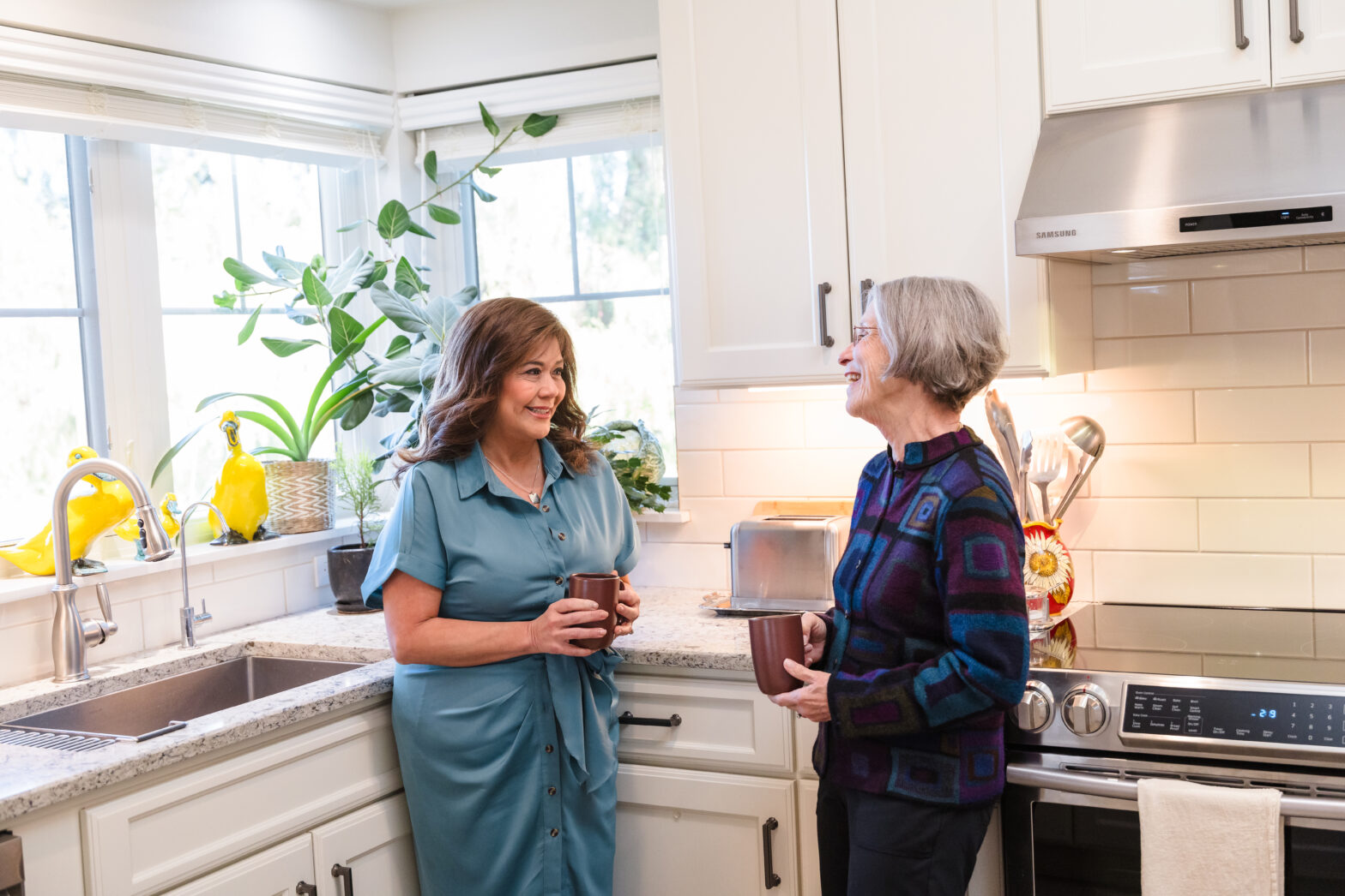 Two senior women stand in the kitchen and talk while holding coffee mugs